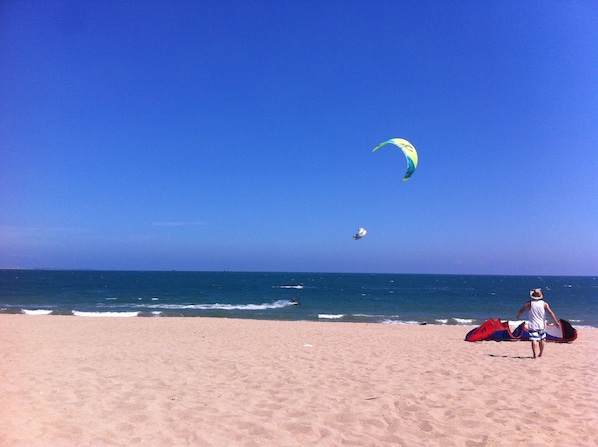 A beach with a clear sky, a person carrying a kite, and the ocean in the background.