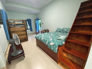 Un dormitorio con cama, escritorio, silla, ventilador y estante de madera.