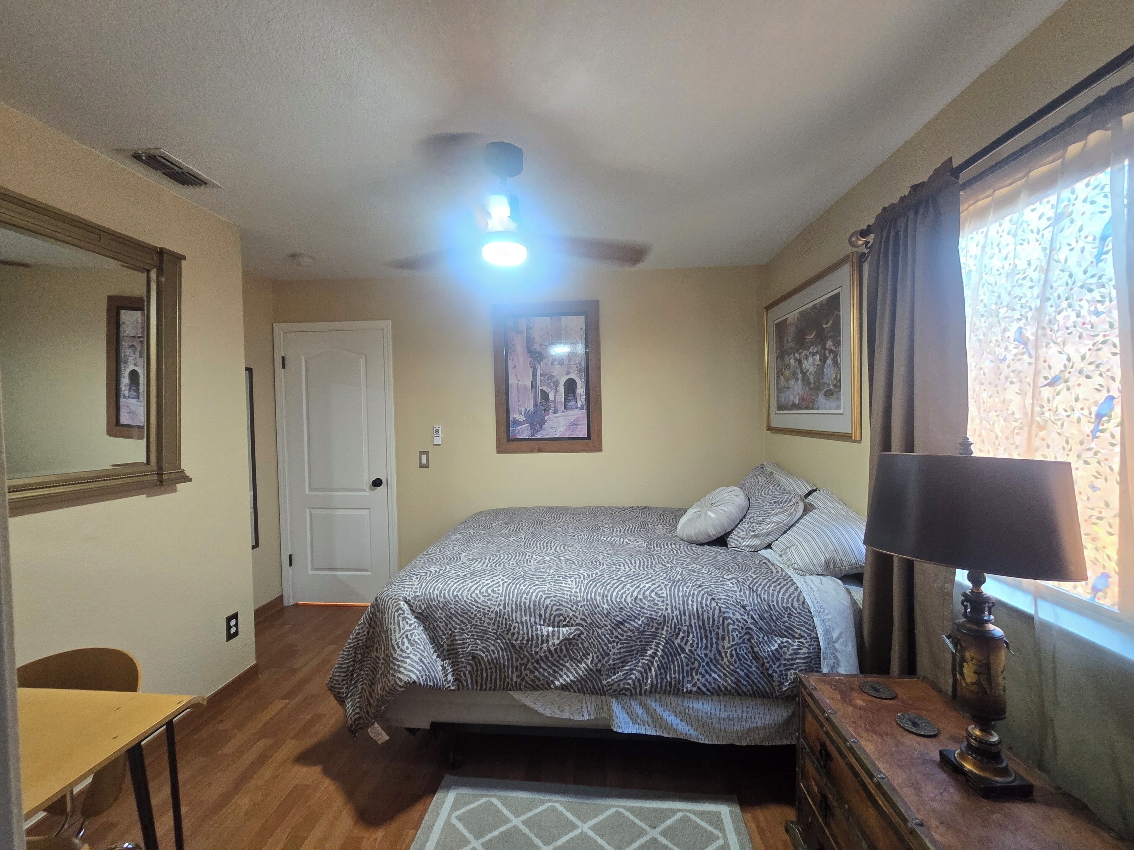 A bedroom with a bed, a nightstand, a lamp, a window with curtains, a door, and a ceiling fan.