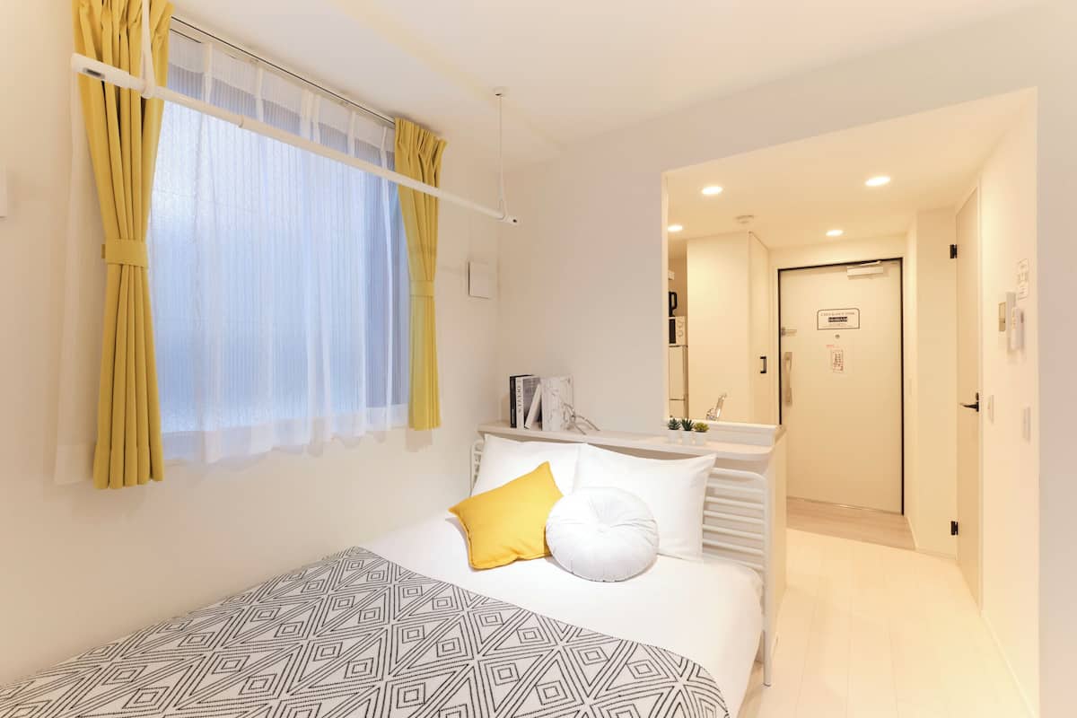Una habitación de hotel con una cama, una mesita de noche, una ventana con cortinas, una puerta y un pasillo.