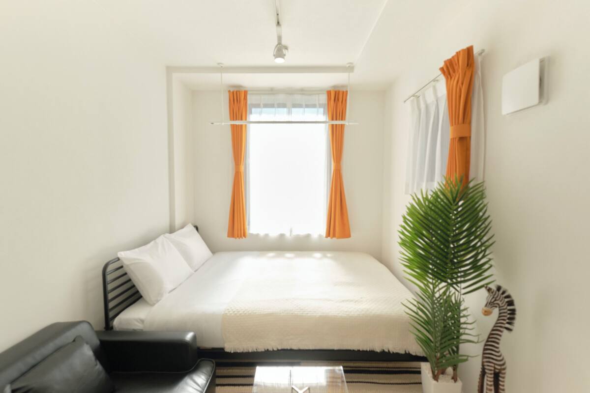Un dormitorio con una cama, un sofá negro, una planta en maceta y cortinas.