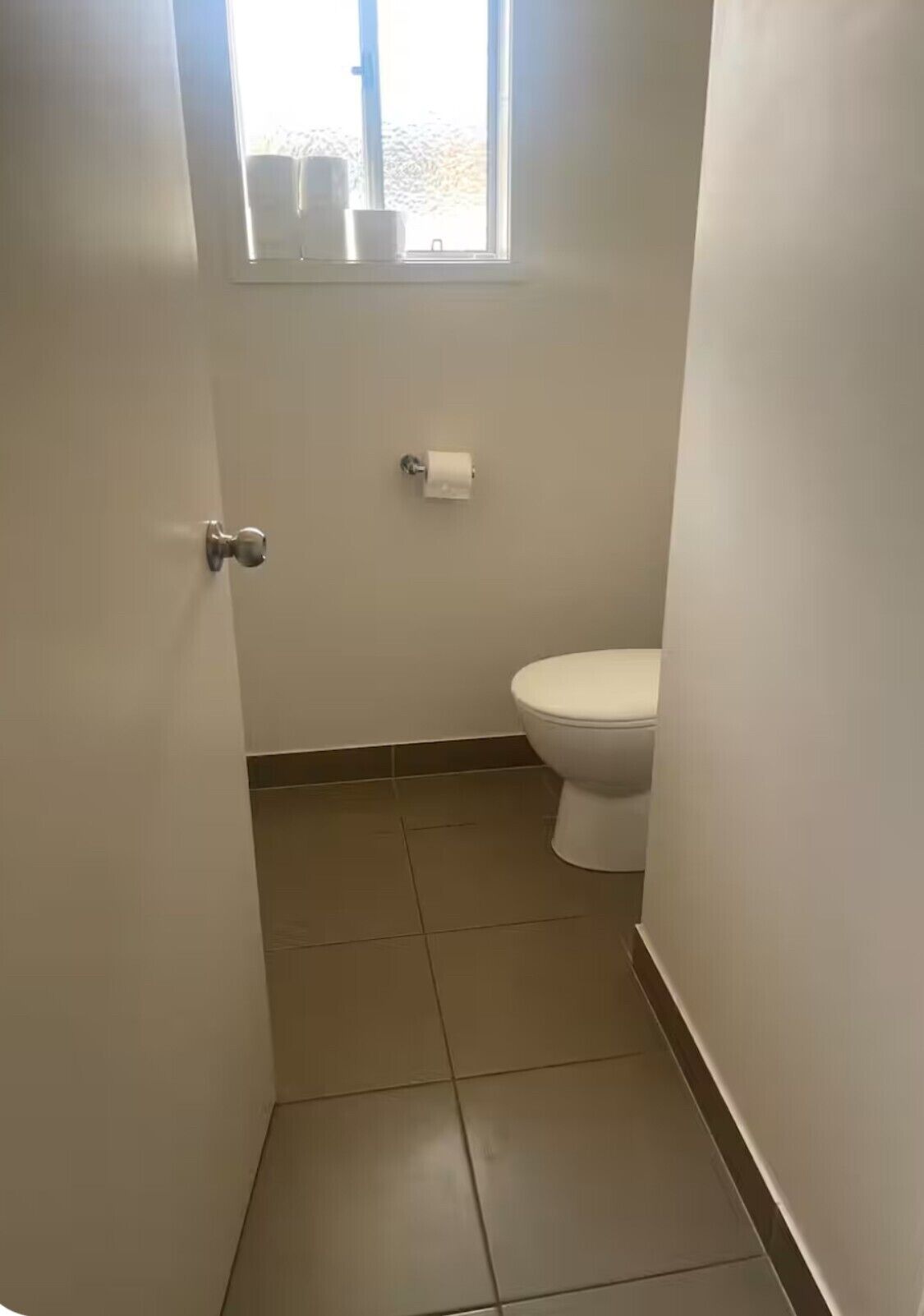 A small, clean bathroom with a toilet, a window, and a roll of toilet paper on the wall.