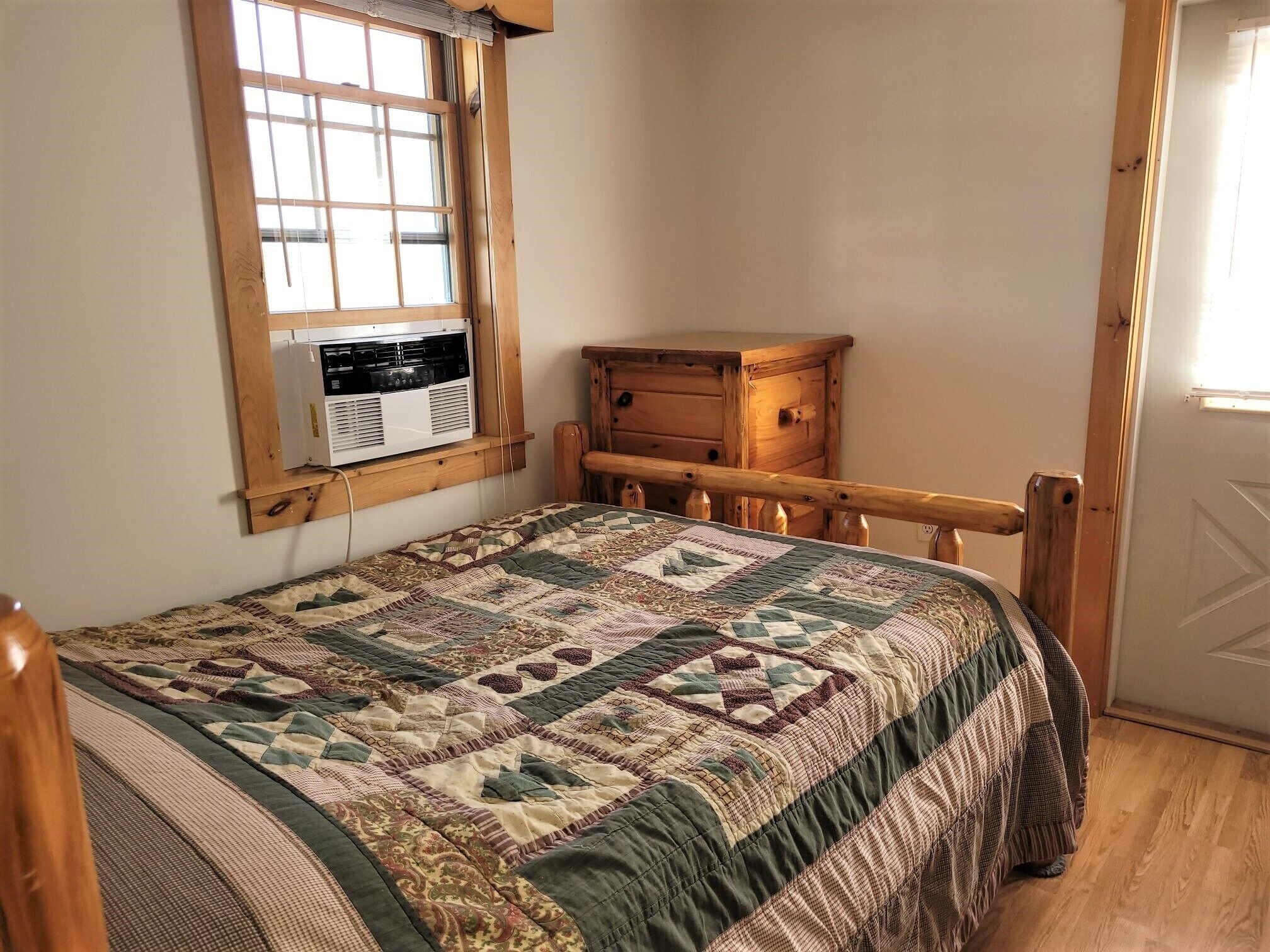 Un dormitorio con una cama de madera, una ventana con aire acondicionado, un aparador de madera y una puerta.