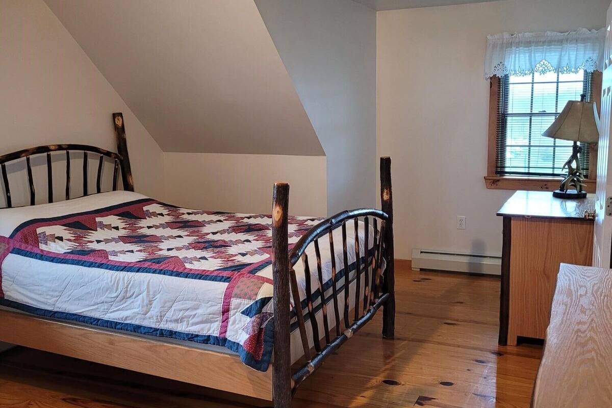 A bedroom with a wooden bed, a quilt, a desk with a lamp, and a window with blinds.