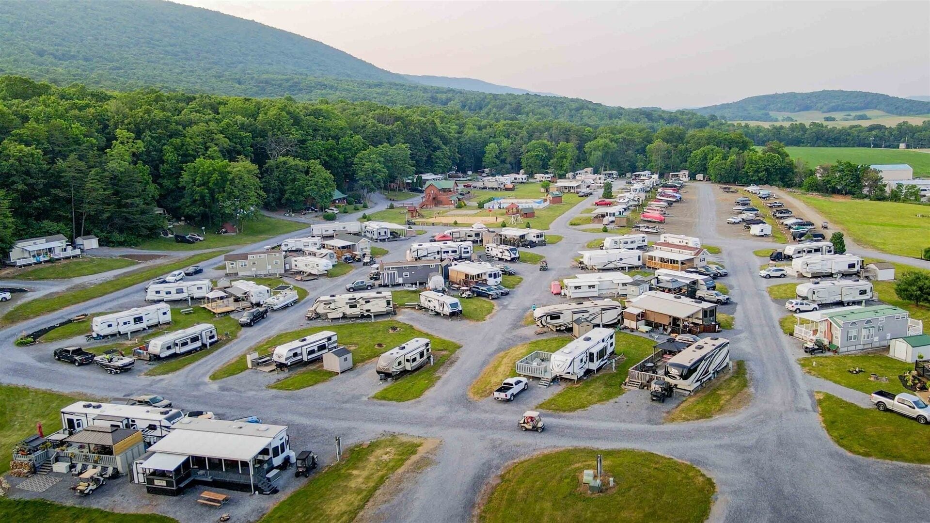 A campground with RVs and trailers parked in organized rows, surrounded by green hills.