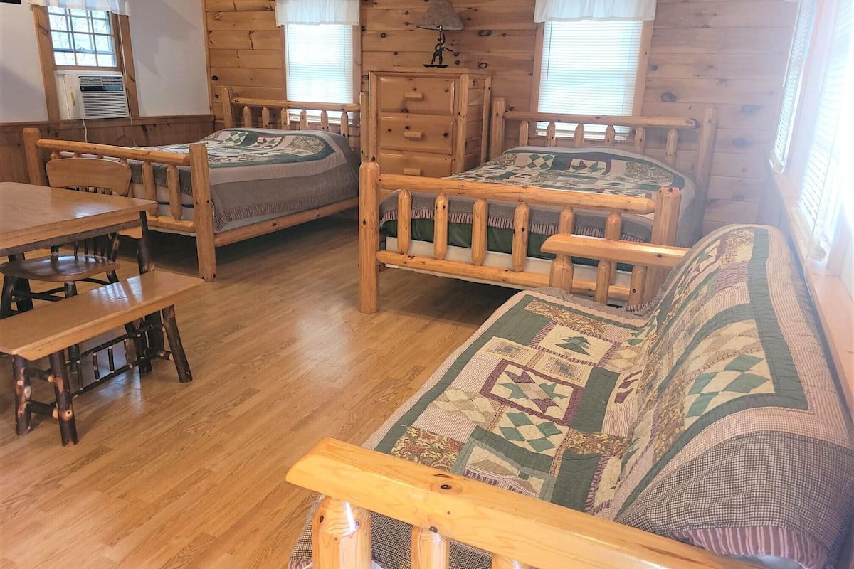 A wooden cabin interior with two beds, a sofa, a dining table, and a dresser.