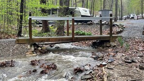 A wooden bridge over a stream with a forested background and RVs in the distance. - James Creek RV Resort (James Creek)