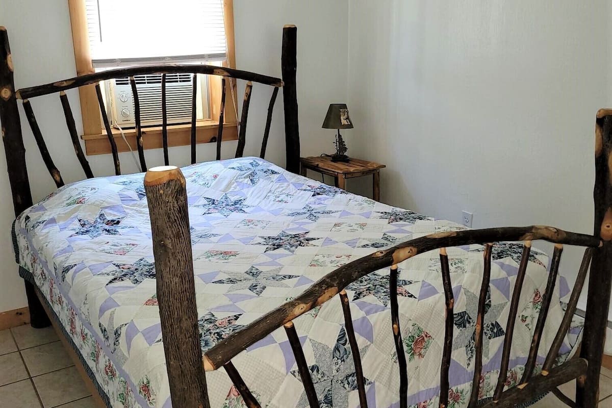 A bedroom with a wooden bed frame, a window with blinds, a bedside table with a lamp, and a floral-patterned bedspread.