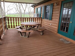 Une terrasse en bois avec une table de pique-nique et des bancs, une véranda et une maison avec une porte et des fenêtres vertes.