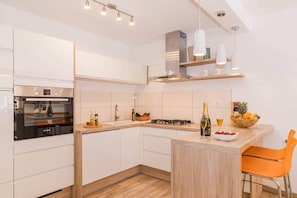 A modern kitchen with white cabinetry, a built-in oven, a gas stove, and a bar area with a bottle of champagne and fruit. - Villa Pietra house situated Galižana (Croatia)