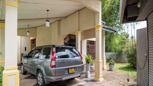 A car parked in a covered parking area with a building entrance, potted plants, and a tiled walkway. - Kethmi Holiday Home (Pannipitiya)