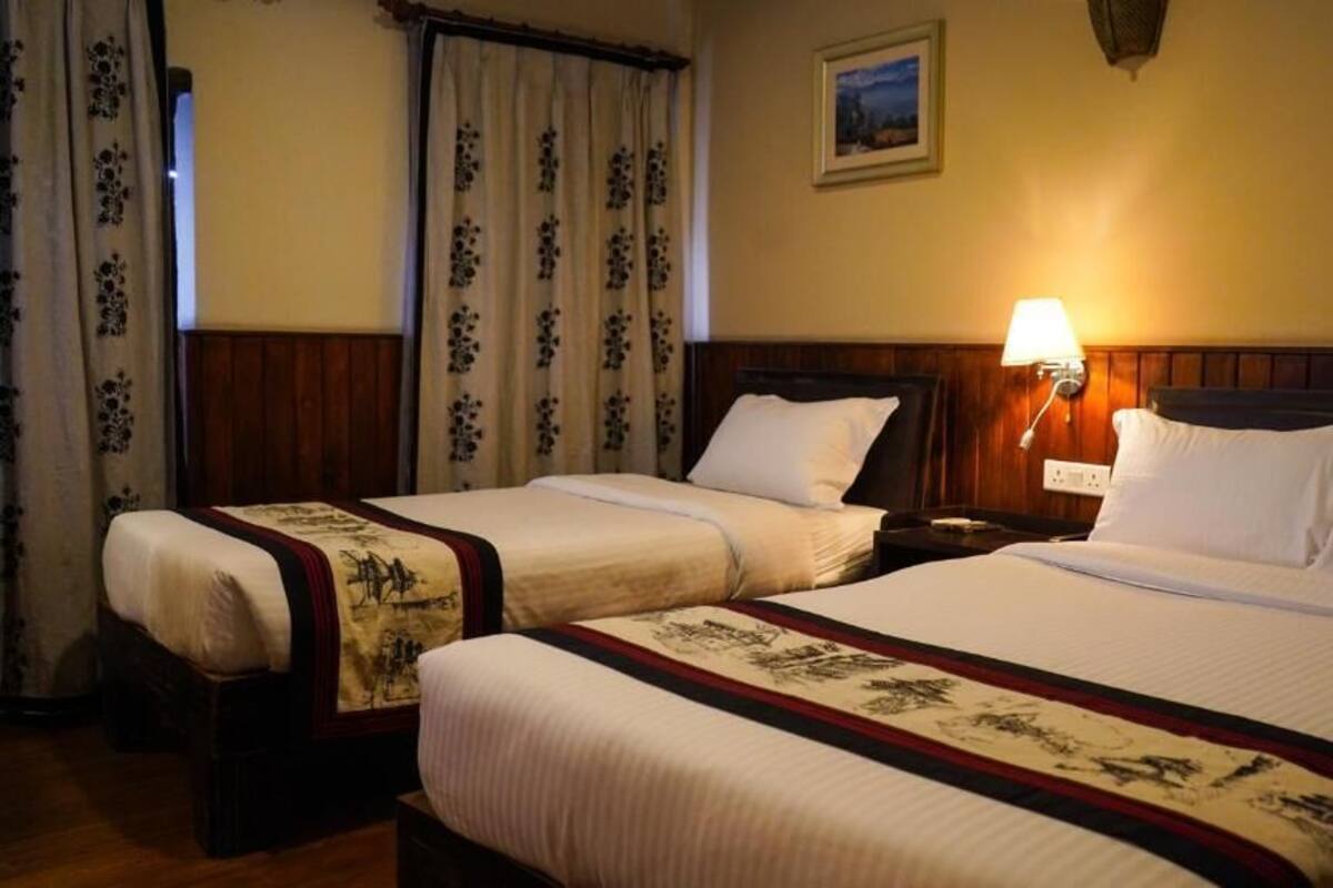 Een hotelkamer met twee bedden, een nachtkastje met een lamp, bloemen gordijnen, en een ingelijst foto aan de muur.