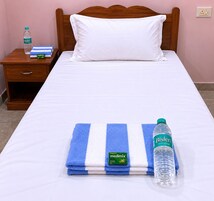 Uma cama bem arrumada com um travesseiro branco, uma toalha listrada em azul e branco e uma garrafa de água Bisleri.