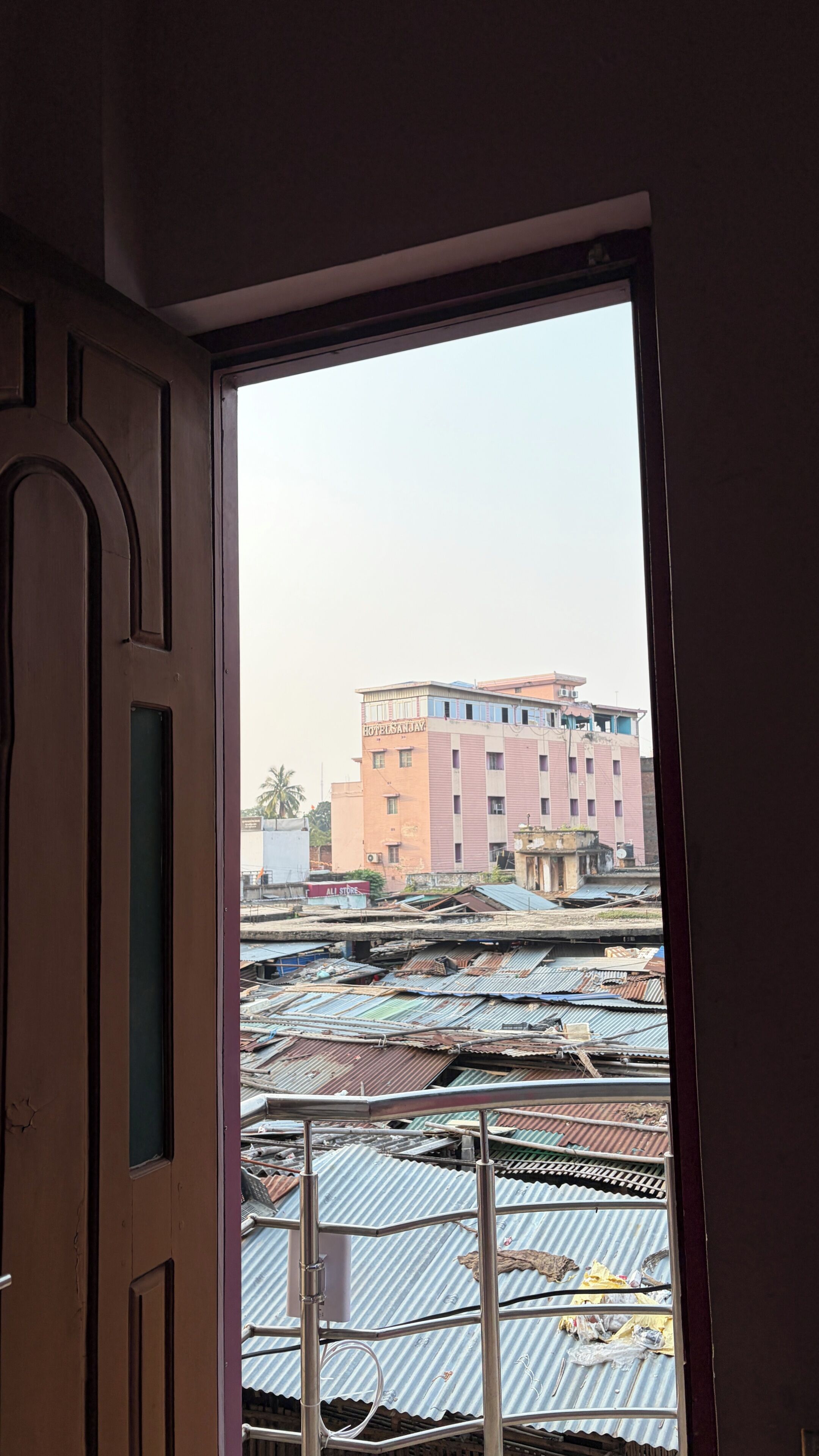 Vista por una ventana de un edificio con un cartel que dice 'HOTEL SANJAY' y una estructura metálica con un cartel que dice 'ALL STORE' debajo.