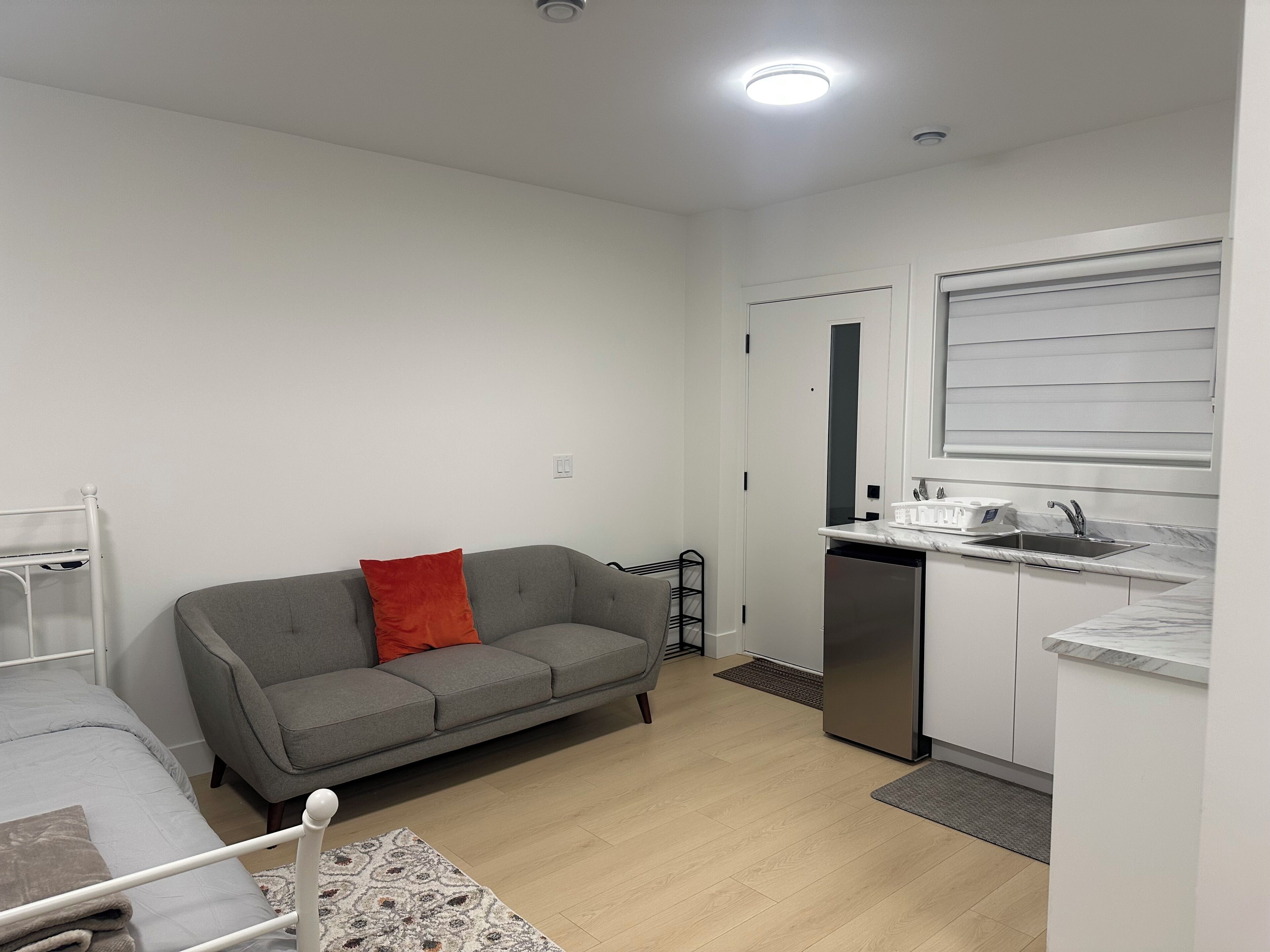 A compact living space with a kitchenette, a grey sofa with a red pillow, a white bed, and a small dining table.. Feel Home out of home