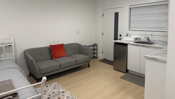 A compact living space with a kitchenette, a grey sofa with a red pillow, a white bed, and a small dining table. - Feel Home out of home (Abbotsford)