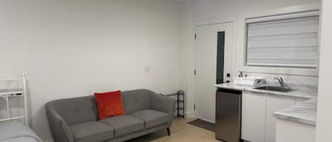 A compact living space with a kitchenette, a grey sofa with a red pillow, a white bed, and a small dining table.