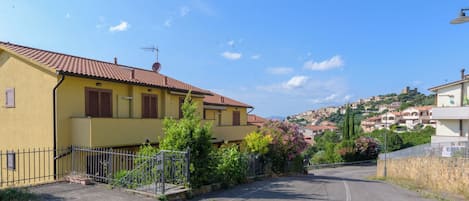 A yellow building with a red-tiled roof, a paved street, and a hilly town in the background.