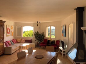 A spacious living room with a sofa, chairs, a dining table, and a TV. There are windows with views of greenery and a fireplace. - House sea view private pool (Bormes-les-Mimosas)