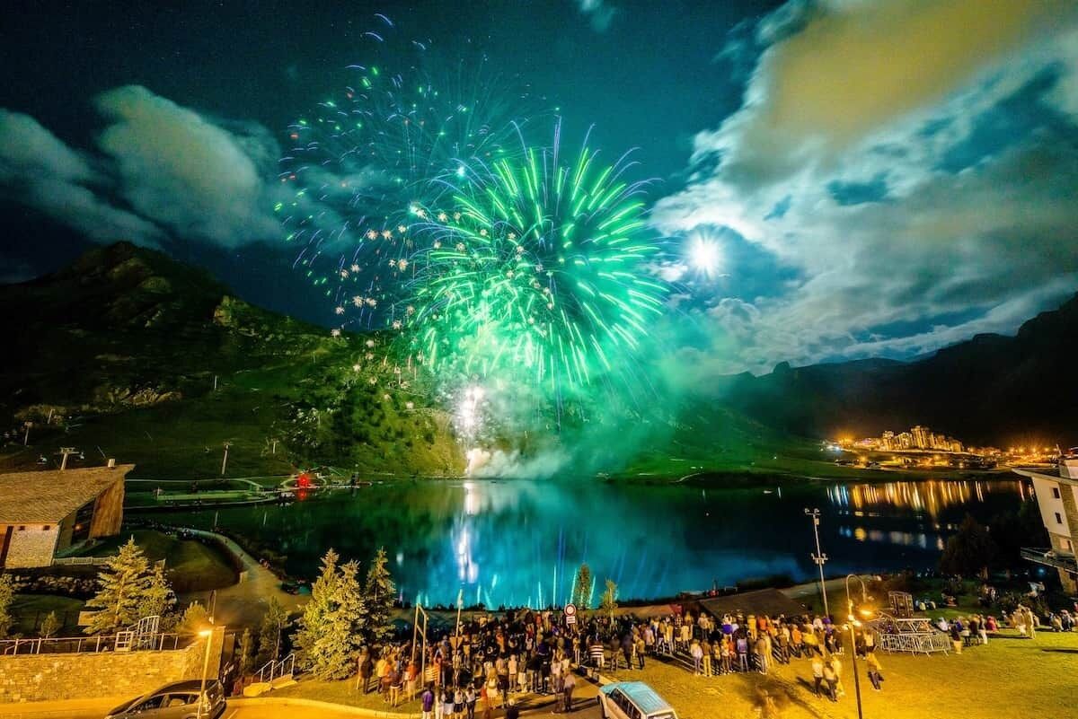 Vista de un lago con fuegos artificiales, una multitud y un edificio de techo inclinado.