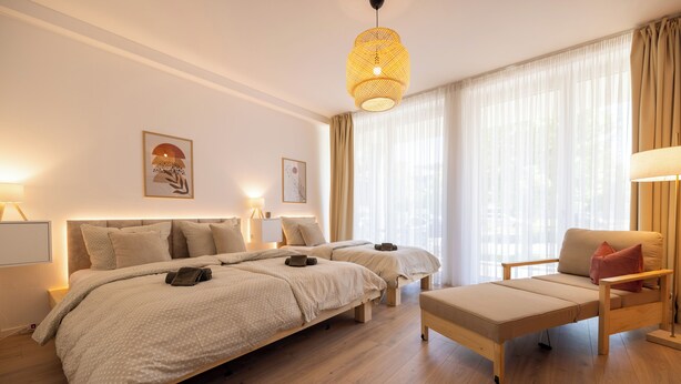 Ein modernes Schlafzimmer mit zwei Betten, einem Schlafsofa, einer Pendelleuchte und großen Fenstern mit transparenten Vorhängen.