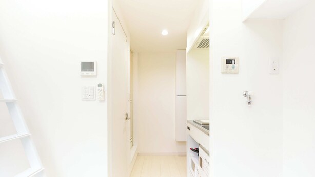 Un pasillo estrecho con una puerta blanca, una pared blanca con un termostato y un estante blanco con un microondas.