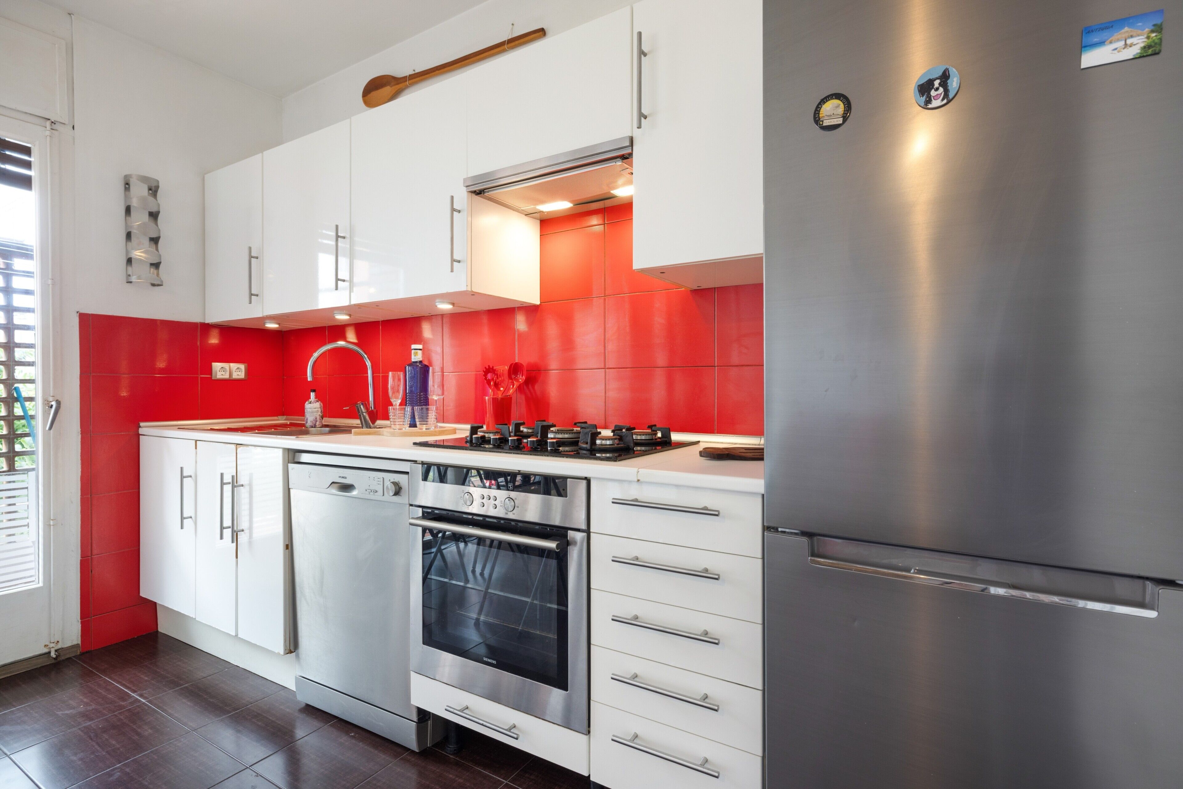 A modern kitchen with white cabinets, a stainless steel refrigerator, a built-in oven, and a red backsplash.
