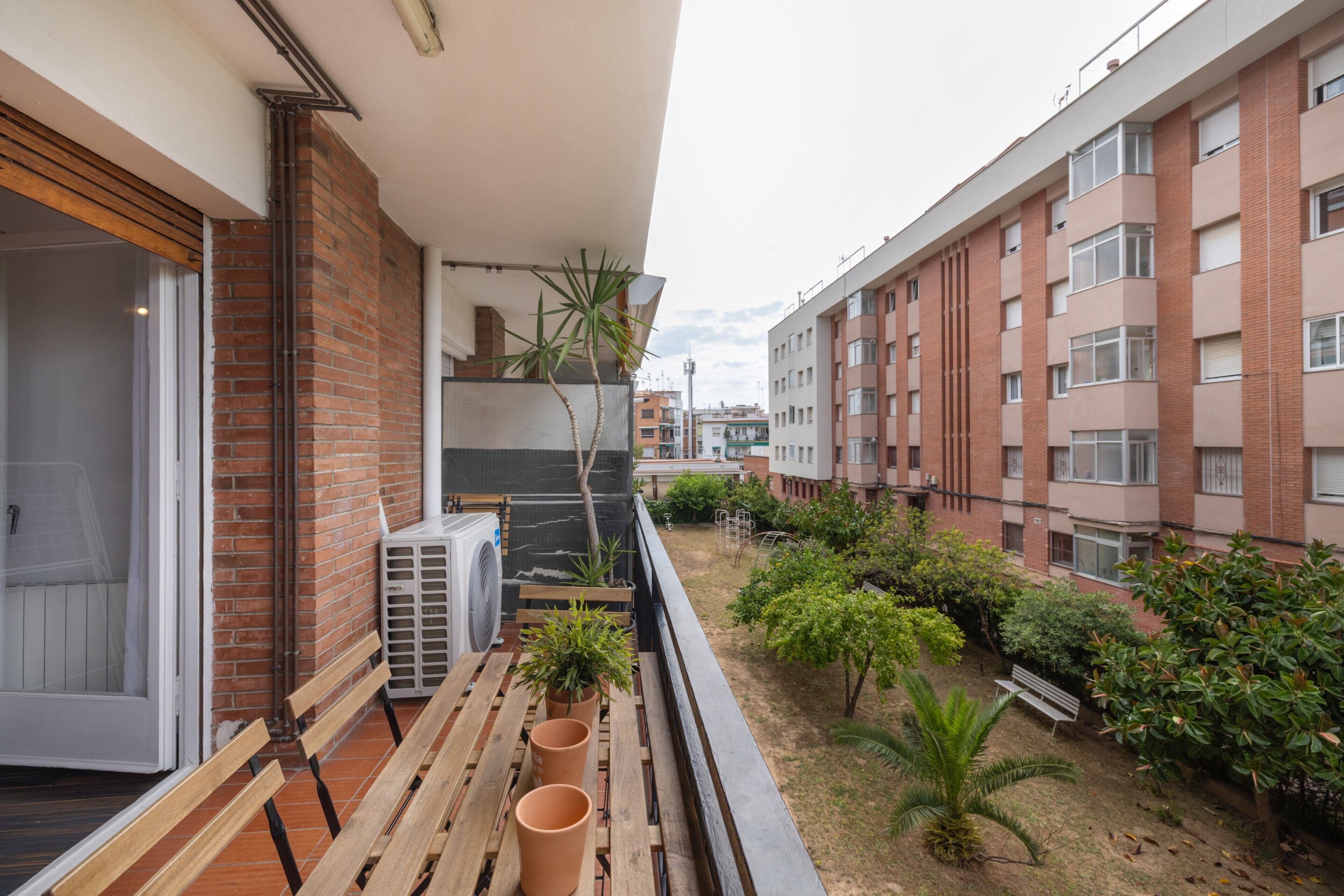 Ein Balkon mit einer hölzernen Sitzbank, Blumentöpfen und Blick auf ein Wohngebiet mit Backsteingebäuden.