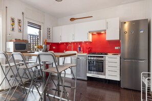 A modern kitchen with white cabinets, a stainless steel refrigerator, a microwave, and a dining area with foldable chairs. - Apartment 'Melody' in Central Sitges with Private Terrace, Wi-Fi, and Air Conditioning (Sitges)