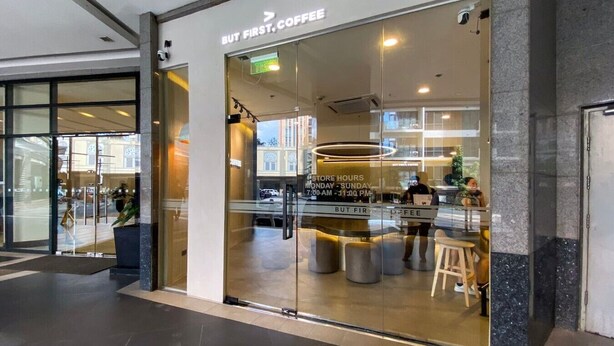 Ett kafé med en glasfasad, en skylt som visar öppettiderna och ett område med sittplatser och barstolar.