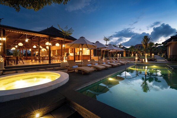 A pool area with lounge chairs and a hot tub, surrounded by a covered outdoor seating area with umbrellas.
