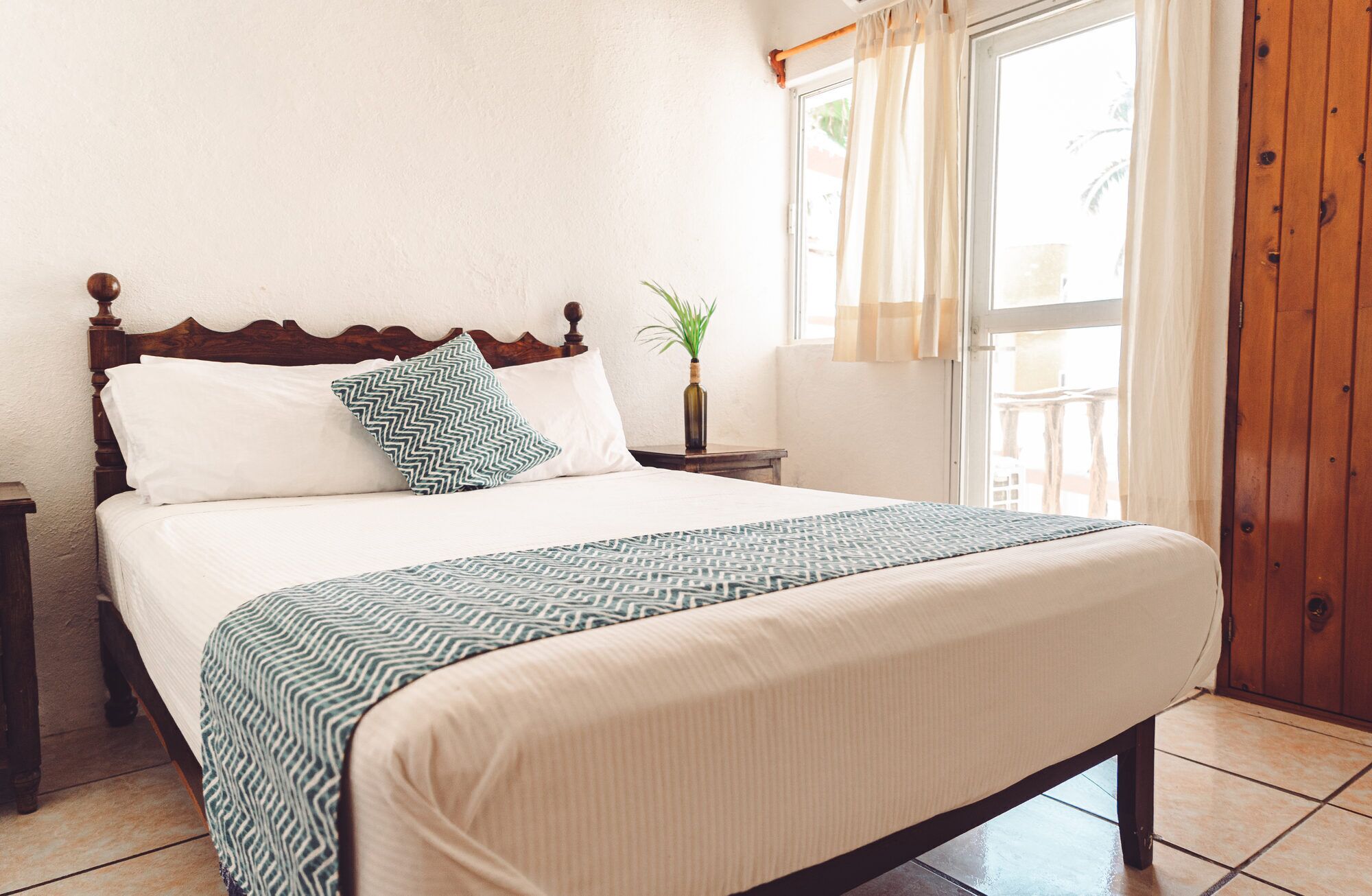 Een netjes opgemaakt bed met een bedsprei en kussens, een nachtkastje met een plant, en een raam met gordijnen.