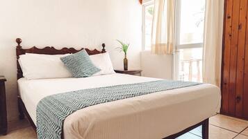 Een netjes opgemaakt bed met een bedsprei en kussens, een nachtkastje met een plant, en een raam met gordijnen.