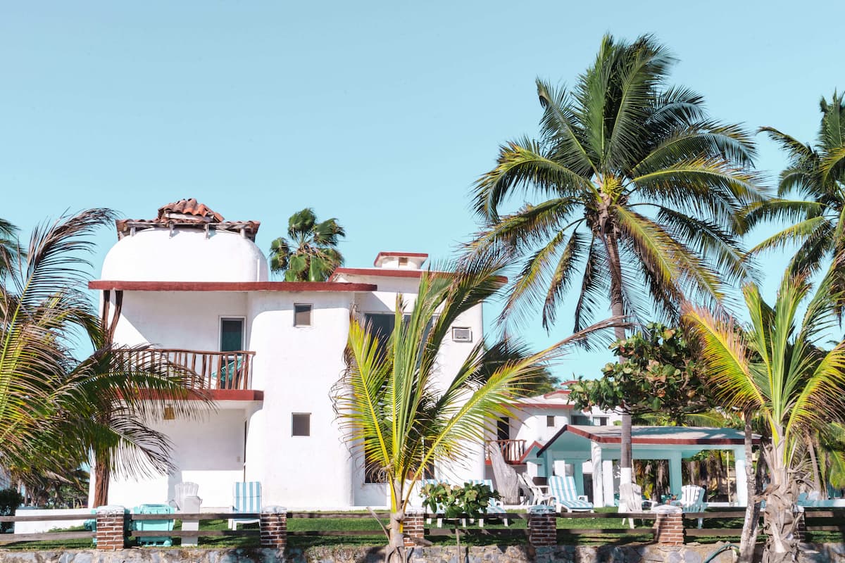 Un edificio blanco con balcón, rodeado de palmeras y un cielo azul despejado.