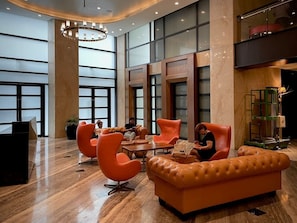A lobby with a modern design, featuring a seating area with orange chairs and a leather sofa, a reception desk, and large windows. - StayHere Fast WiFi 5 Star View in Makati (Makati)