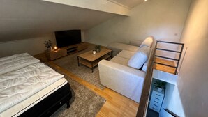 Ein kompakter Wohnbereich mit Bett, Sofa, Couchtisch, Fernseher und einer kleinen Treppe.