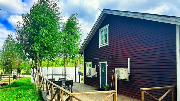 Eine Holzterrasse mit Tisch und Stühlen, ein dunkel gefärbtes Gebäude mit Tür und Fenstern sowie ein umzäunter Bereich mit Grünpflanzen.
