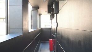 A narrow corridor with a red trash bin, a window on the left, and a wall-mounted speaker on the right. - Stylewood Nishi-Shinjuku 2 (Shibuya)