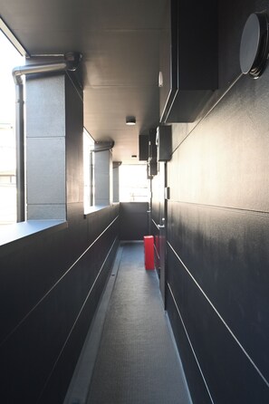 A narrow corridor with a red trash bin, a window on the left, and a wall-mounted speaker on the right. - Stylewood Nishi-Shinjuku 2 (Shibuya)