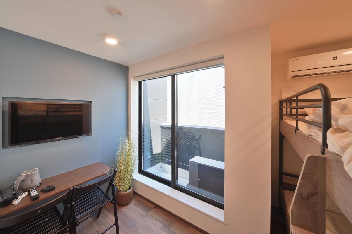 A compact hotel room with a bed, a TV, a desk with a chair, a potted plant, and a large window offering a view of the outside.