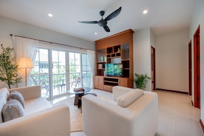 A modern living room with a ceiling fan, a TV on a wooden entertainment unit, and a balcony with a table and chairs.