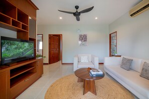 A living room with a TV, wooden entertainment unit, white sofa, and a ceiling fan.