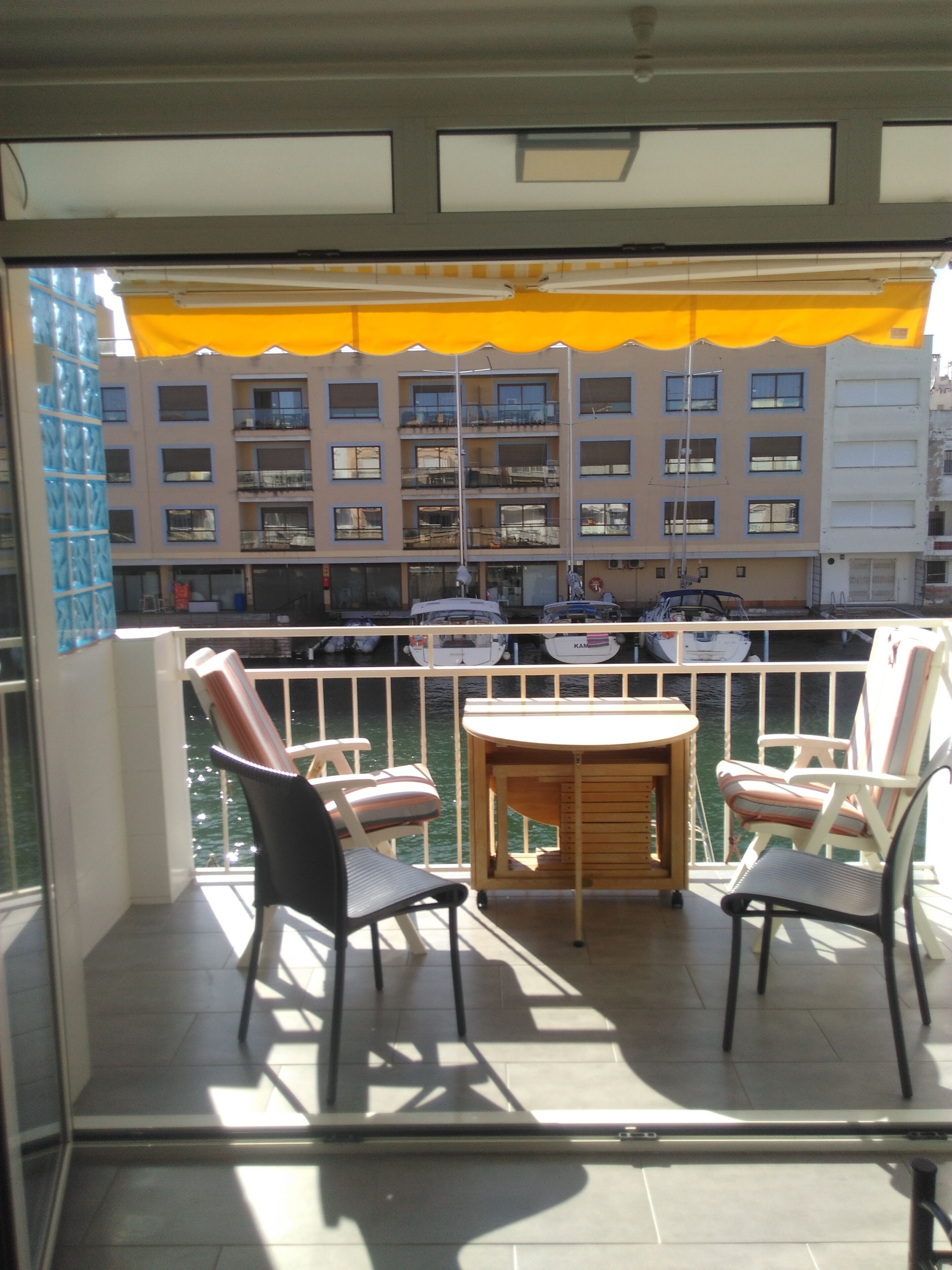 Un balcon avec deux chaises et une table, donnant sur une marina où sont amarrés des bateaux, ainsi que sur un immeuble doté de nombreux balcons.
