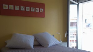 Een bed met een gewatteerde spreideken, een rode muur met ingelijste foto’s en een raam met uitzicht op gebouwen.