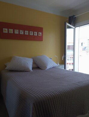 A bed with a quilted cover, a red wall with framed pictures, and a window with a view of buildings. - Apartment 2 bedrooms 1 Living room with balcony (Empuriabrava)