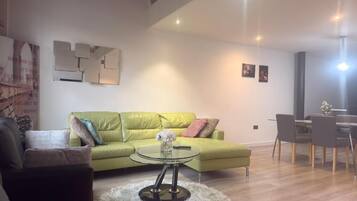 A modern living room with a green sofa, a glass coffee table, and a dining area with chairs.