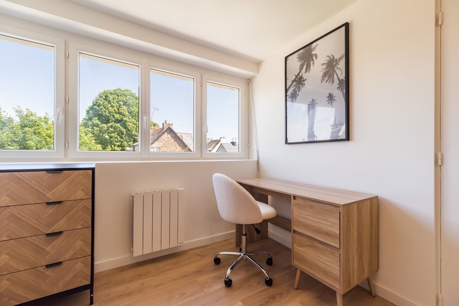 Une pièce avec un bureau en bois, une chaise blanche, un radiateur et une fenêtre donnant sur des arbres et des immeubles.