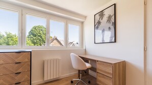 A room with a wooden desk, a white chair, a radiator, and a window with a view of trees and buildings.
