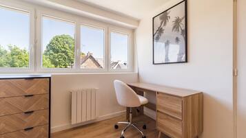 Une pièce avec un bureau en bois, une chaise blanche, un radiateur et une fenêtre donnant sur des arbres et des immeubles.
