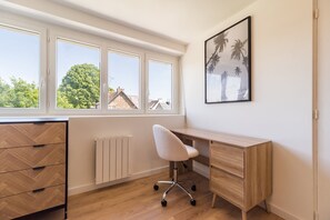 A room with a wooden desk, a white chair, a radiator, and a window with a view of trees and buildings. - Cozy loft in the heart of Guingamp (Guingamp)